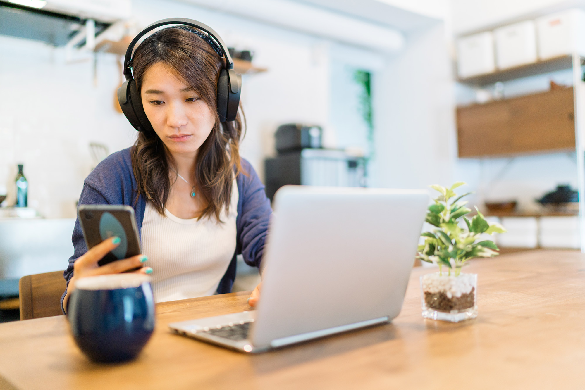 A woman with headphones sits at a desk in front of a laptop looking at her phone