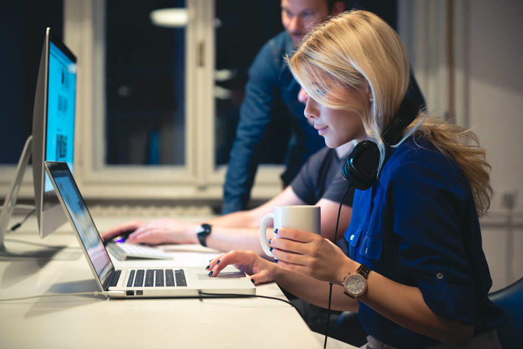 Woman typing on laptop with headphones on ears and her coworkers are defocused in background