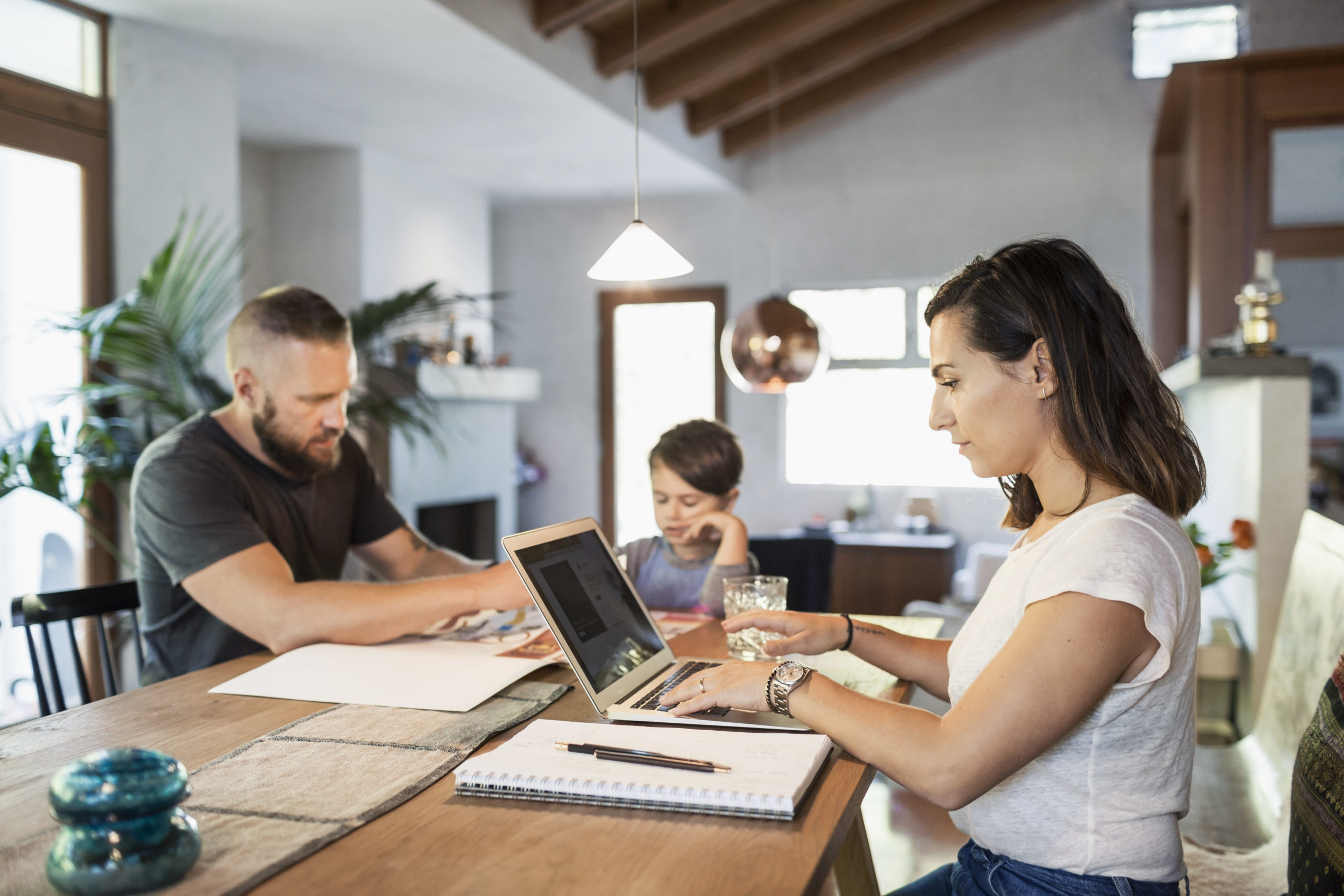 Woman working on laptop at dining table with family in background