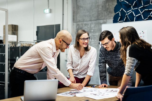 A team of four business people is standing in front a desk in a bright office room. They ambitiously point at documents while smiling and looking at the charts and notes.