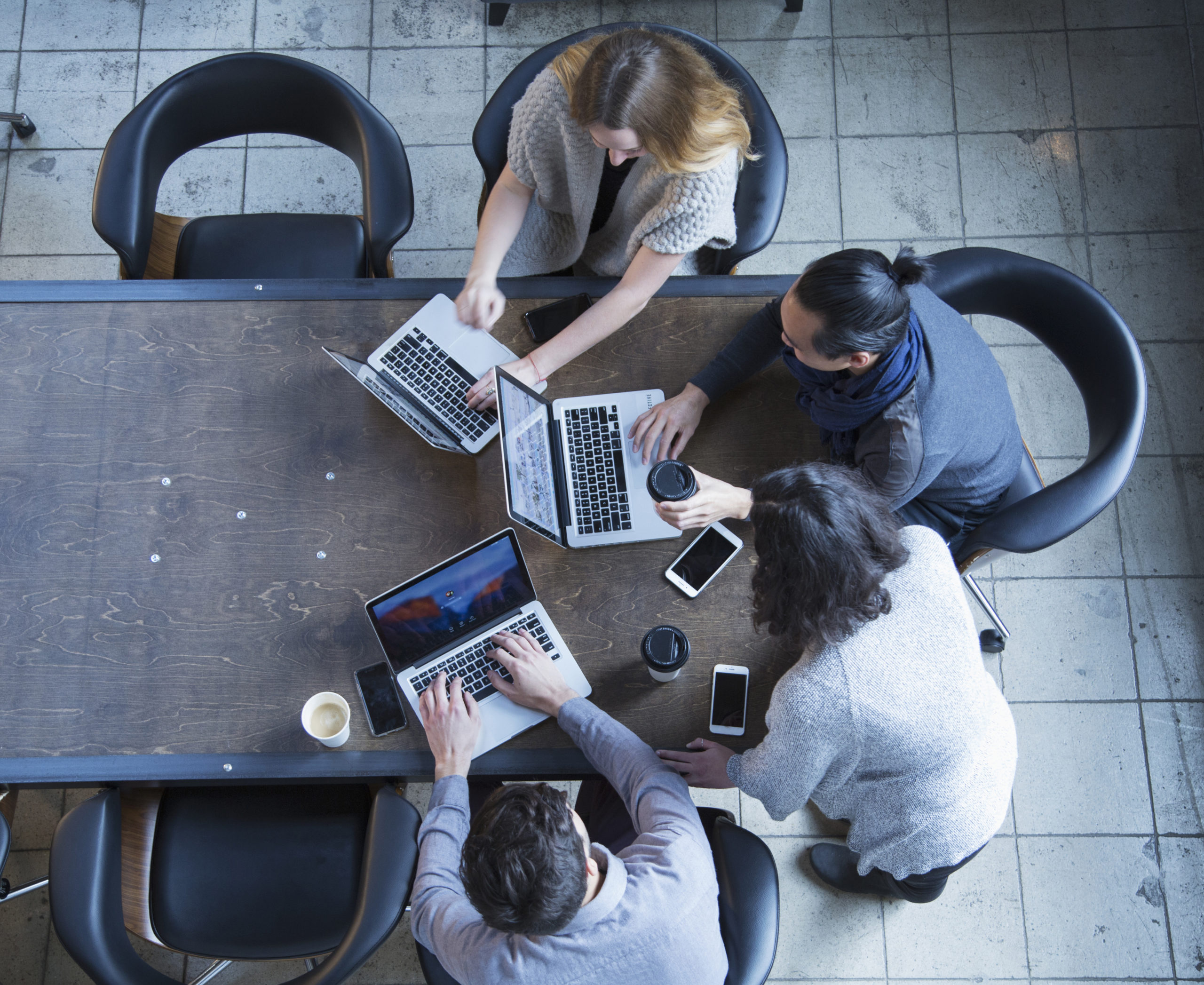Aerial view of a team of four people huddled around three laptops at the end of a table.