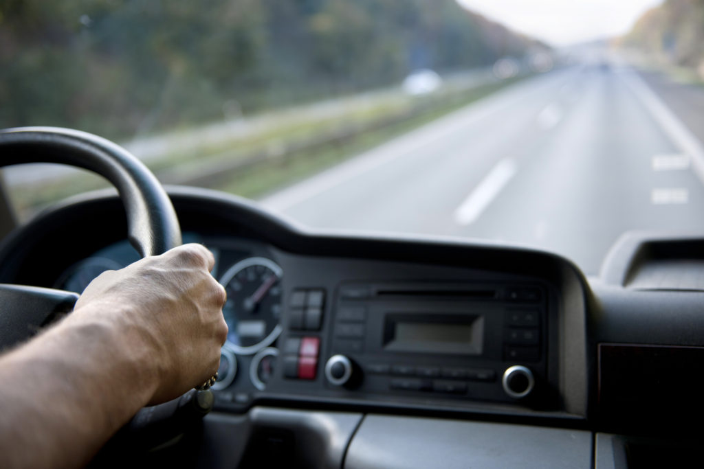 A driver's hands on the steering wheel of a car with a clear, two-lane road ahead of them.