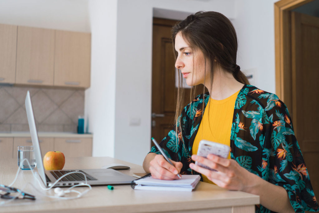 Woman sitting at desk looking at computer and writing things down in notepad