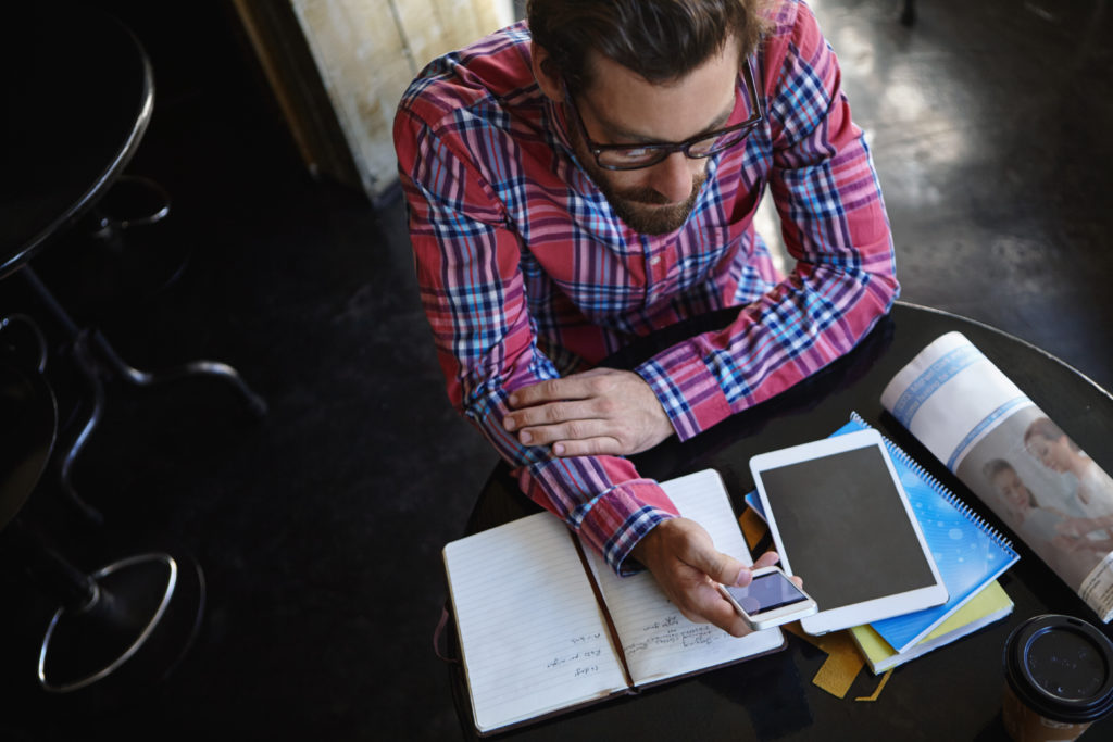 Shot of a man sitting at a table in a cafe with his cellphone and tablet in front of him