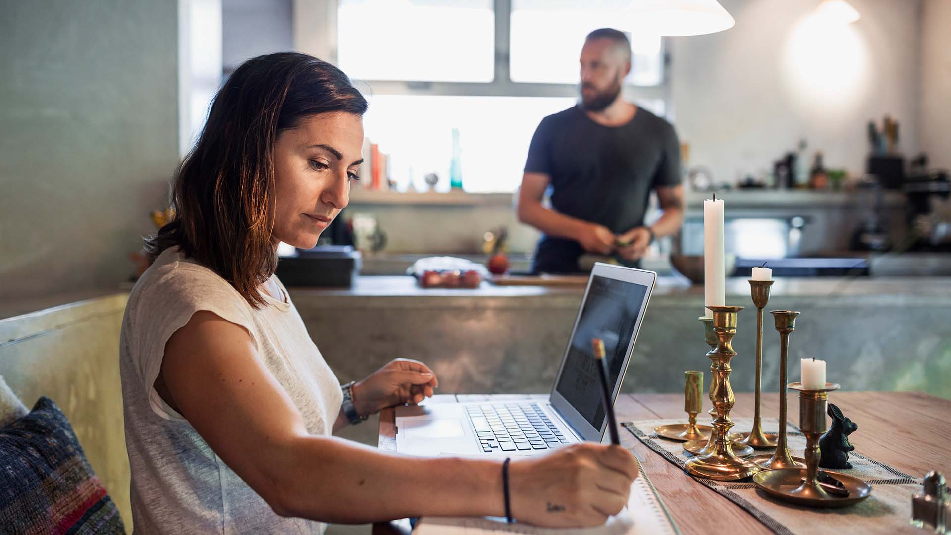 Woman works at home desk with man behind her