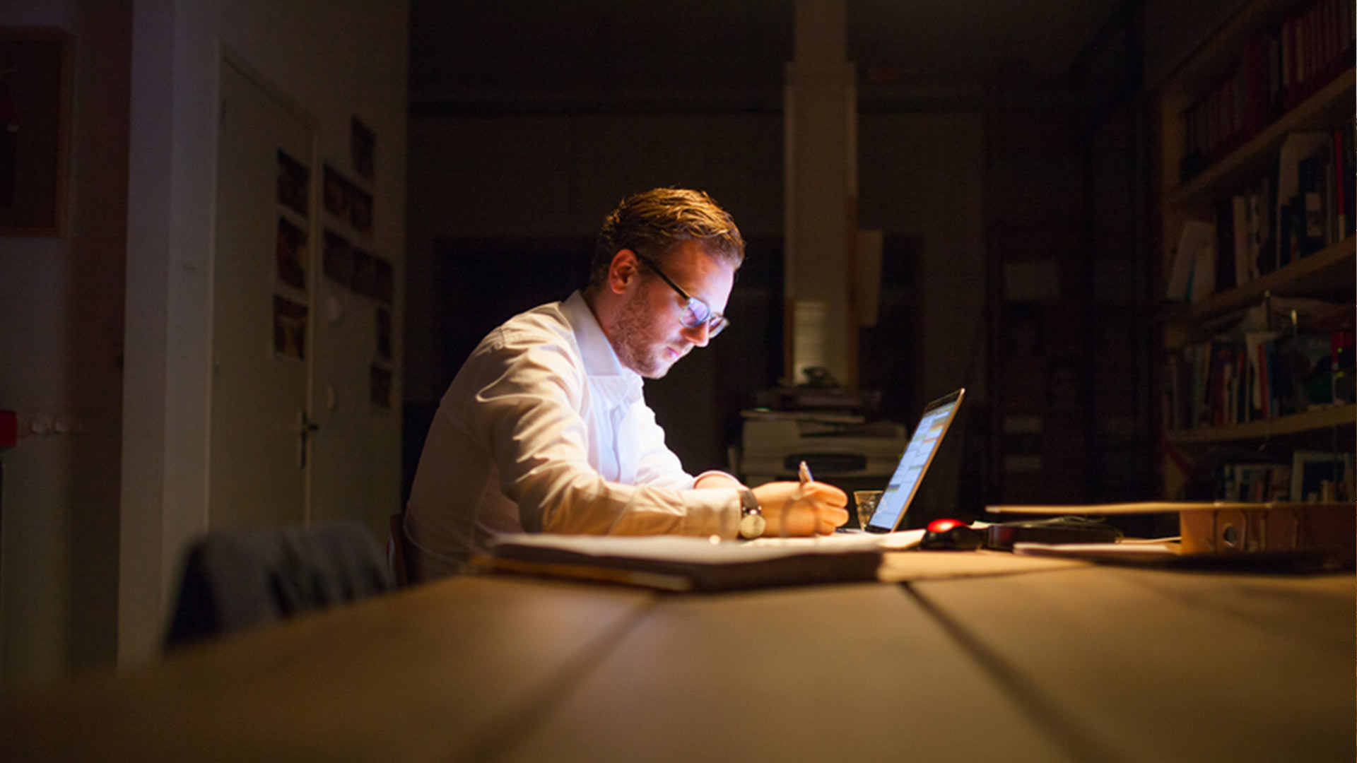 man in dark looking at computer writing on notepad