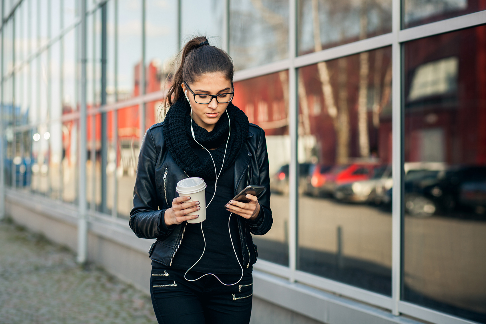 Woman in headphones walking down the street looking at phone and holding coffee