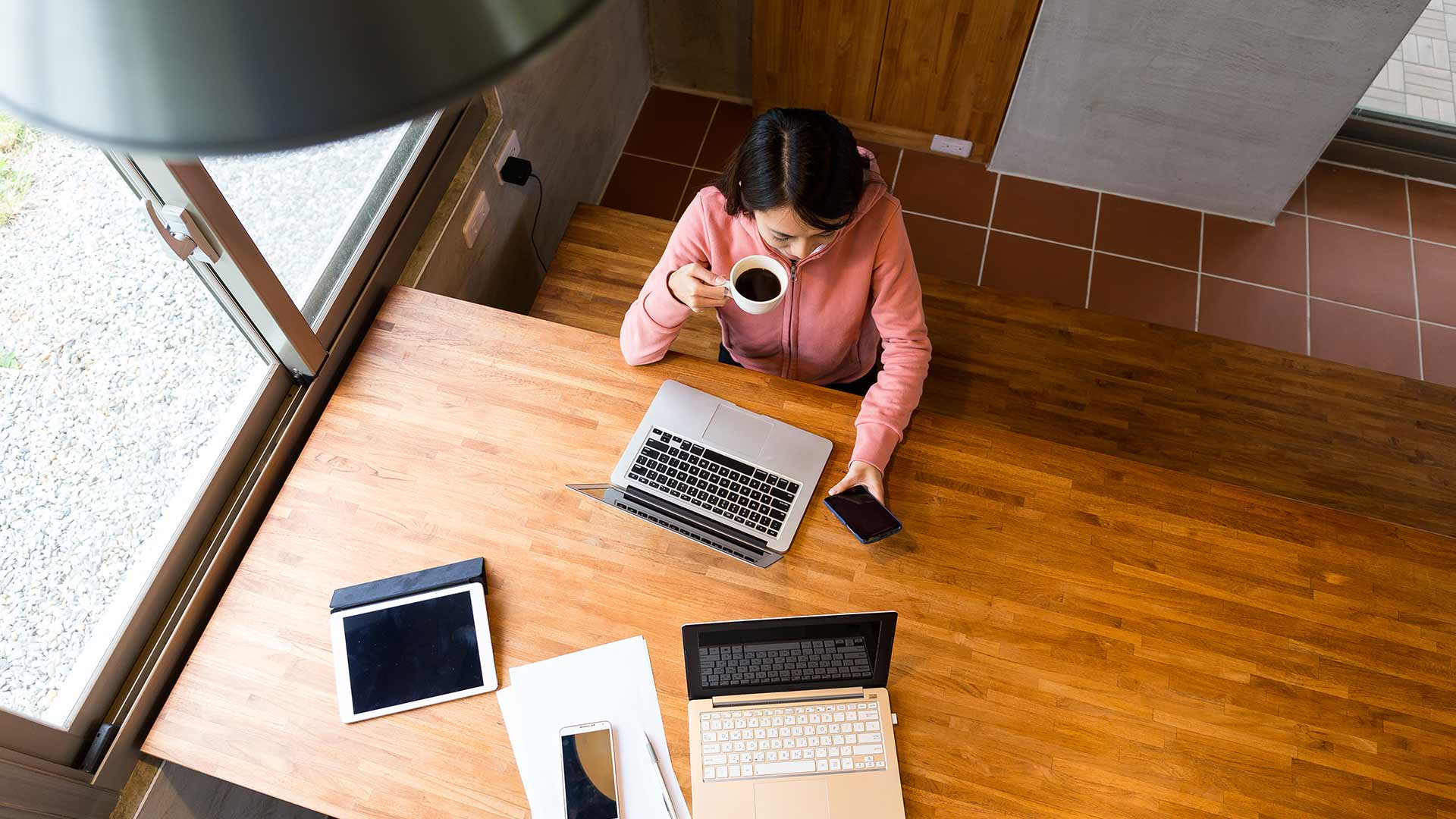 Woman at desk drinking coffee with several screens laid out in front of her. She is looking at her phone