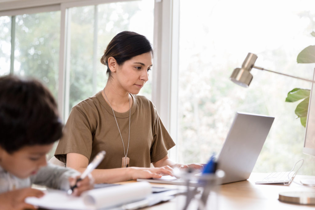 Woman concentrates while using a laptop in her home office while her son is drawing a picture in the foreground.