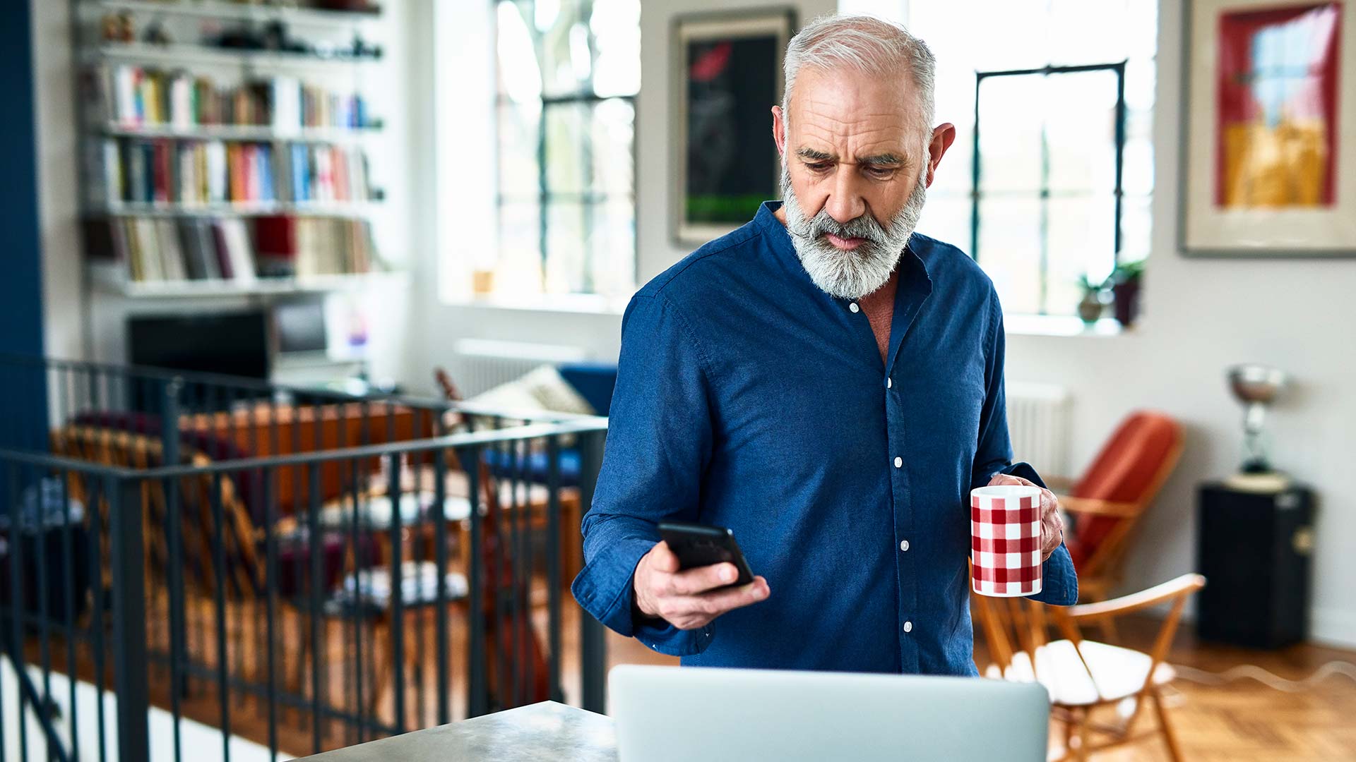 Man in home looking at his phone while holding a cup of coffee.