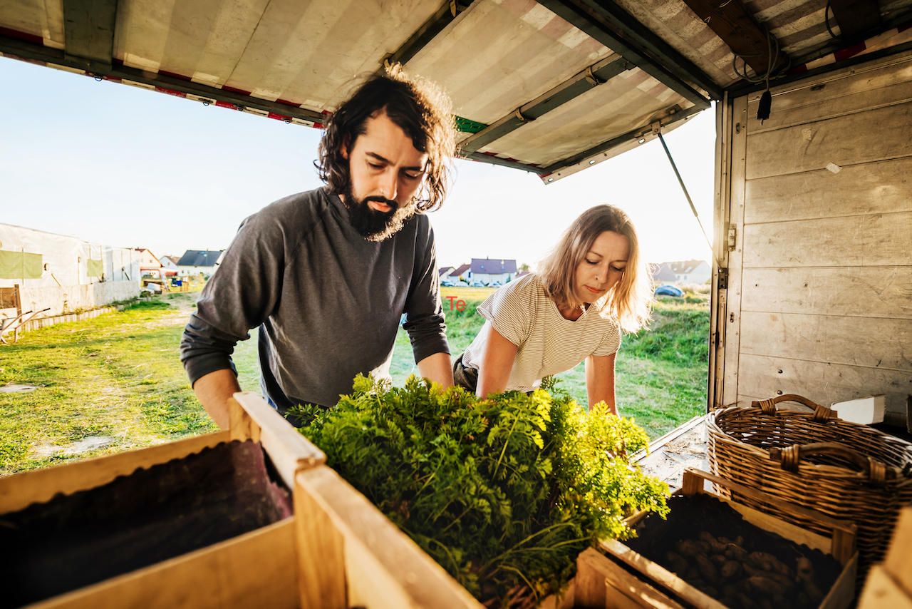 Two farmers are loading crates of various freshly harvested goods onto a truck