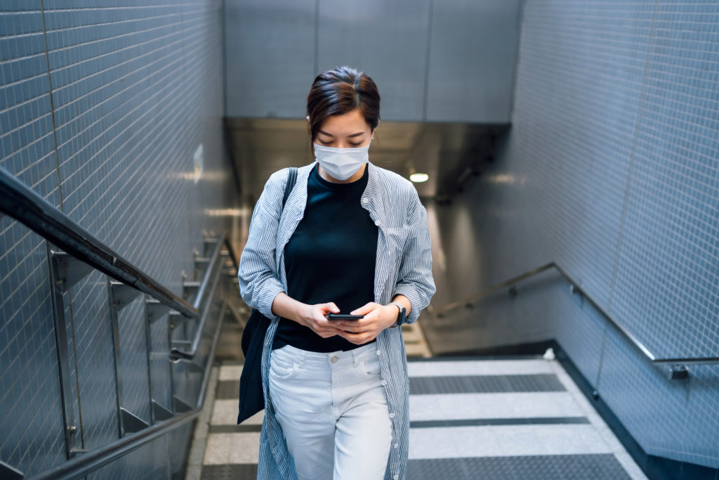 Businesswoman with protective face mask using smartphone, while walking upstairs in the subway station. 