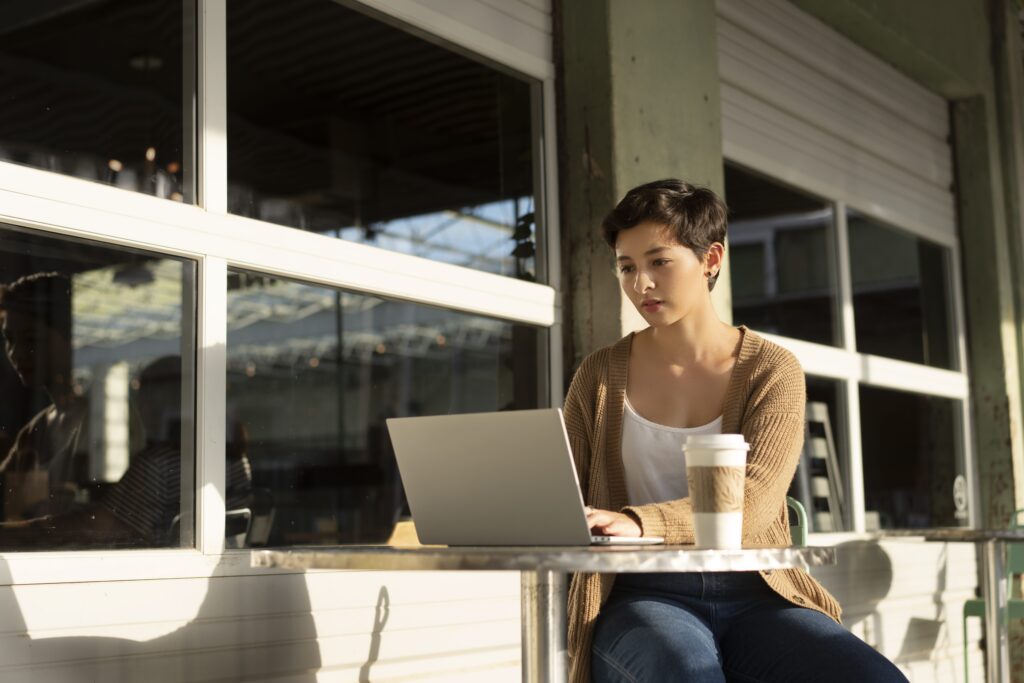 Person working on a laptop outside a cafe with windows in the background and a coffee cup on the table