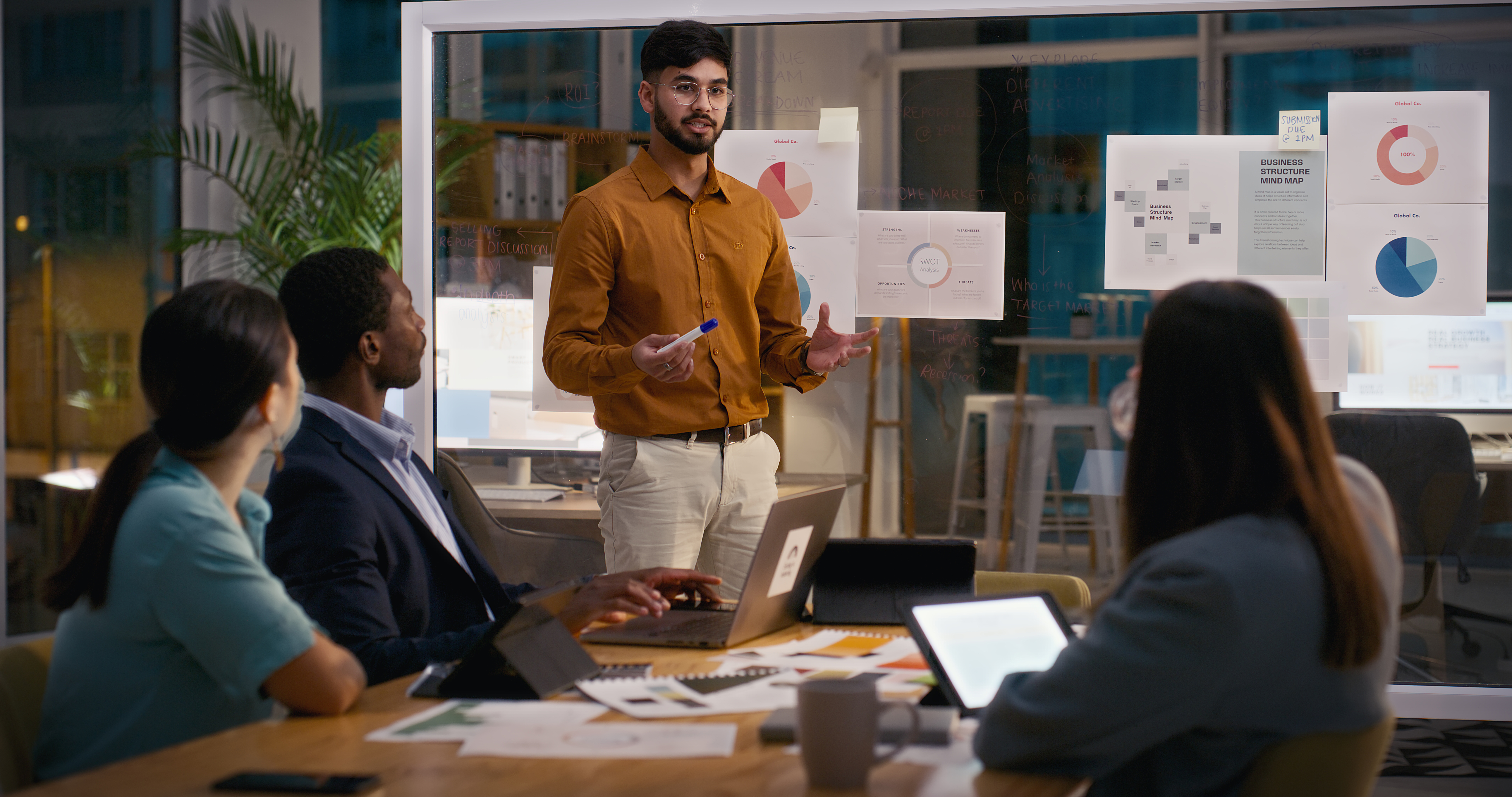 Colleagues meeting in the office with man standing in front of standing charts while seated people around him look on