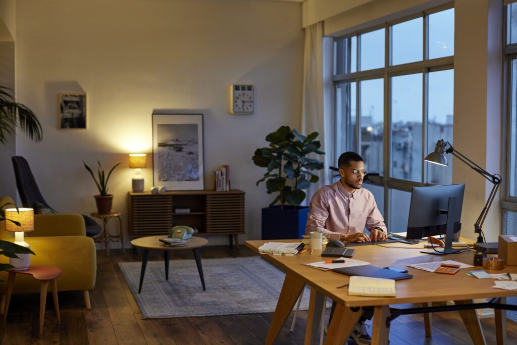 Young professional using computer at table in apartment. Entrepreneur is working in home office in the evening with living room in the background