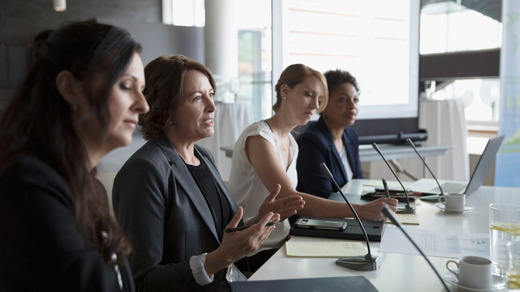 Four women on a panel with one speaking and one taking notes.