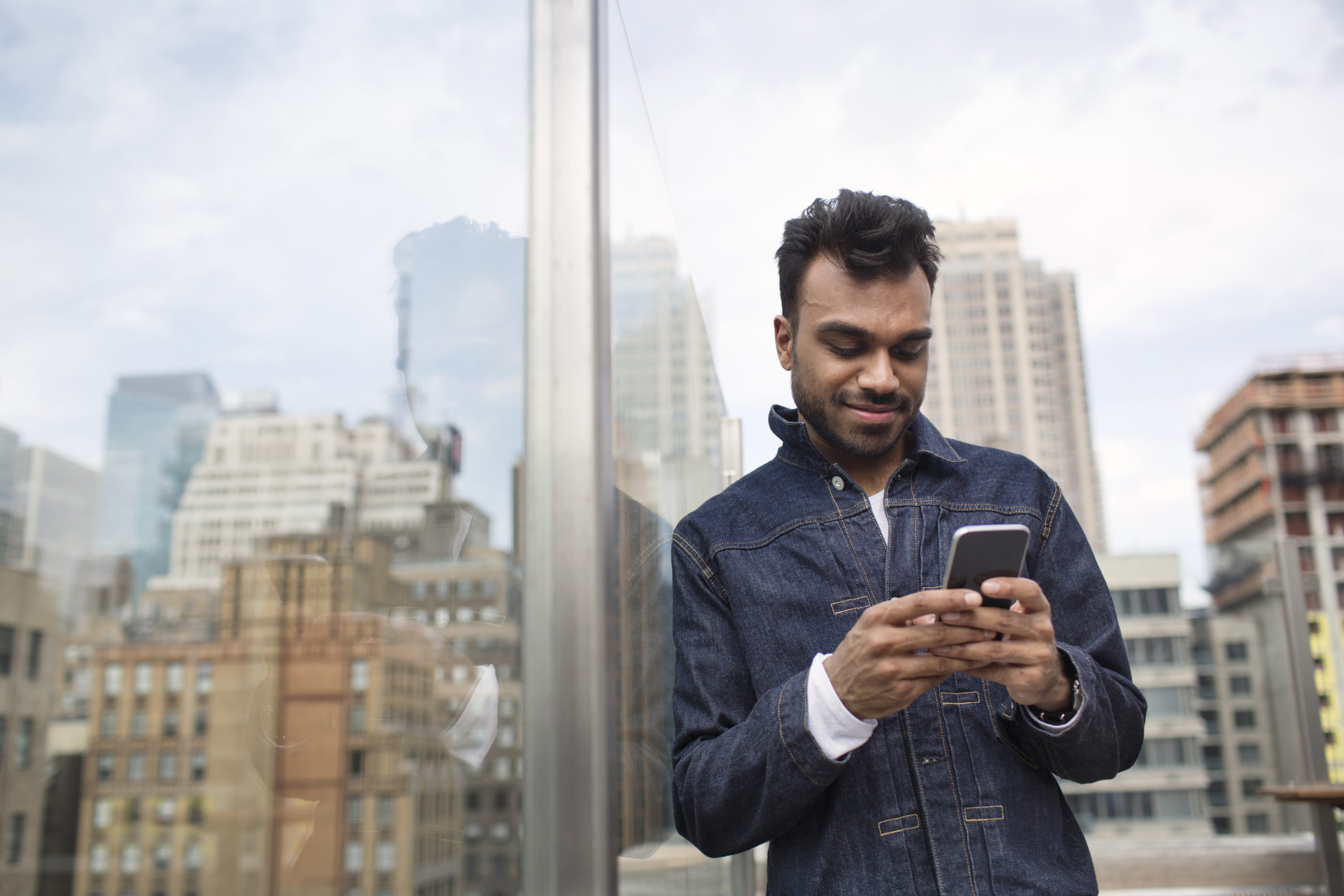 Smiling young man using phone while standing by window at rooftop restaurant.