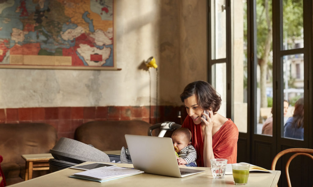 Mother carrying baby using phone while looking in laptop at table in restaurant