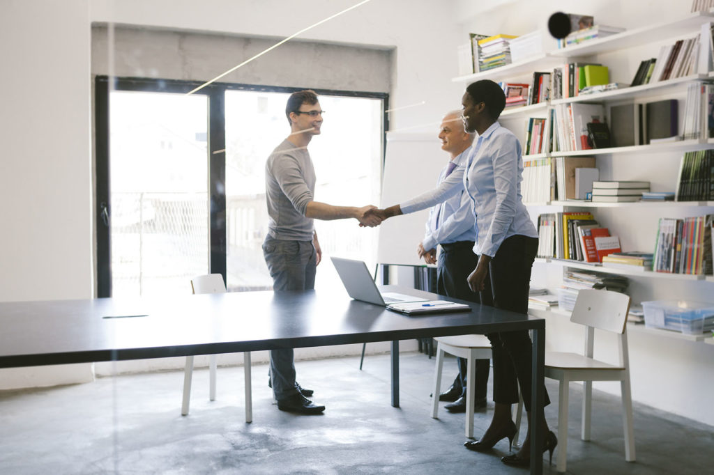 Job Interview with young male candidate in modern office. He is coming into interview room and handshake with mature businessman and african businesswoman.