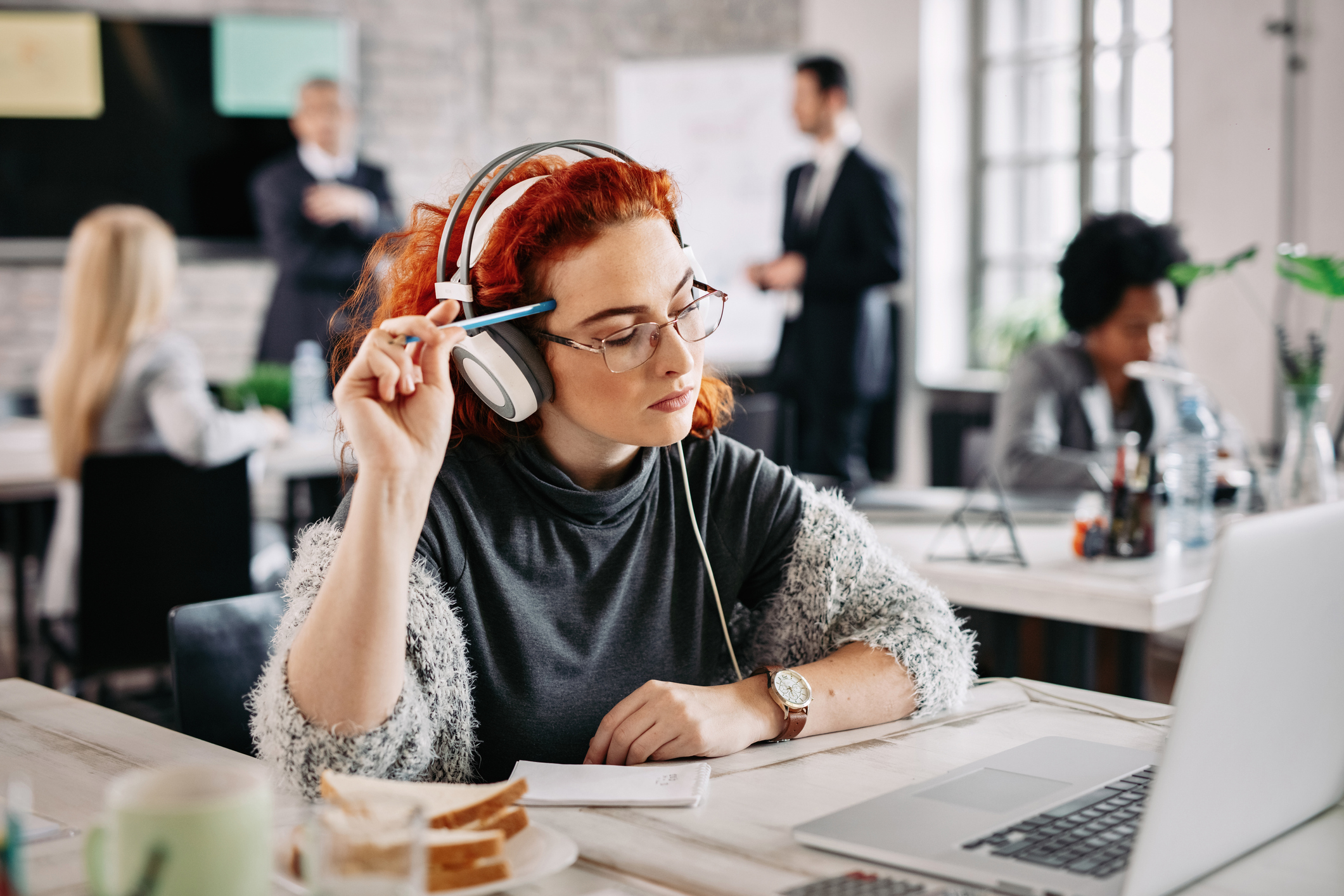 Young female entrepreneur using laptop and thinking about e-mail she has received while listening music on headphones at work. There are people in the background.