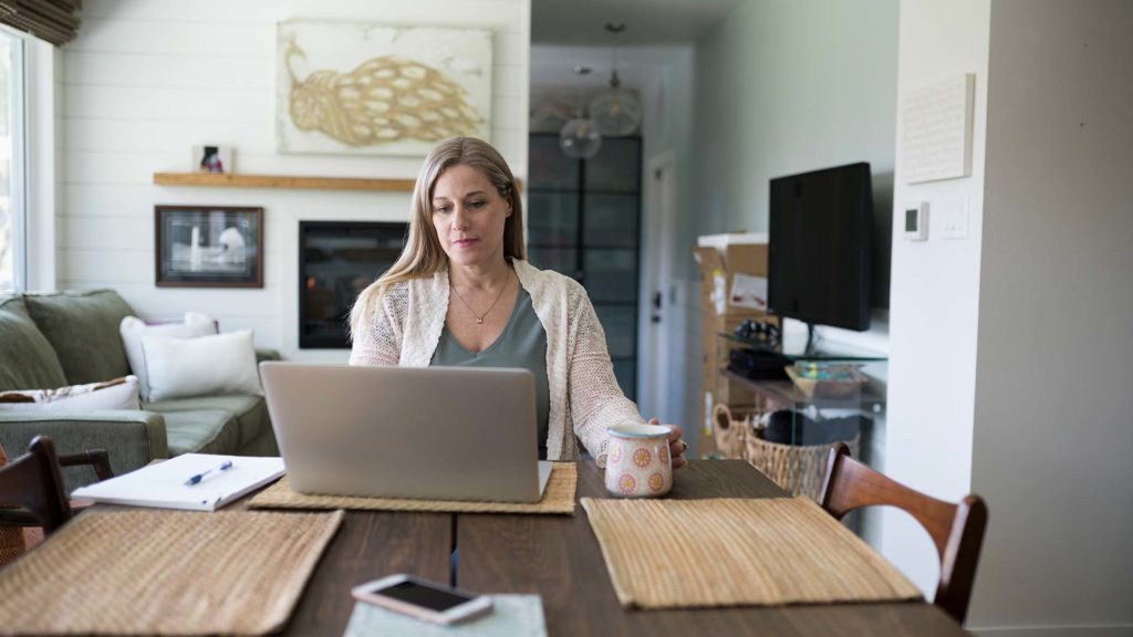 A woman working from home at her dining room table in front of her laptop with a mug in her hand.