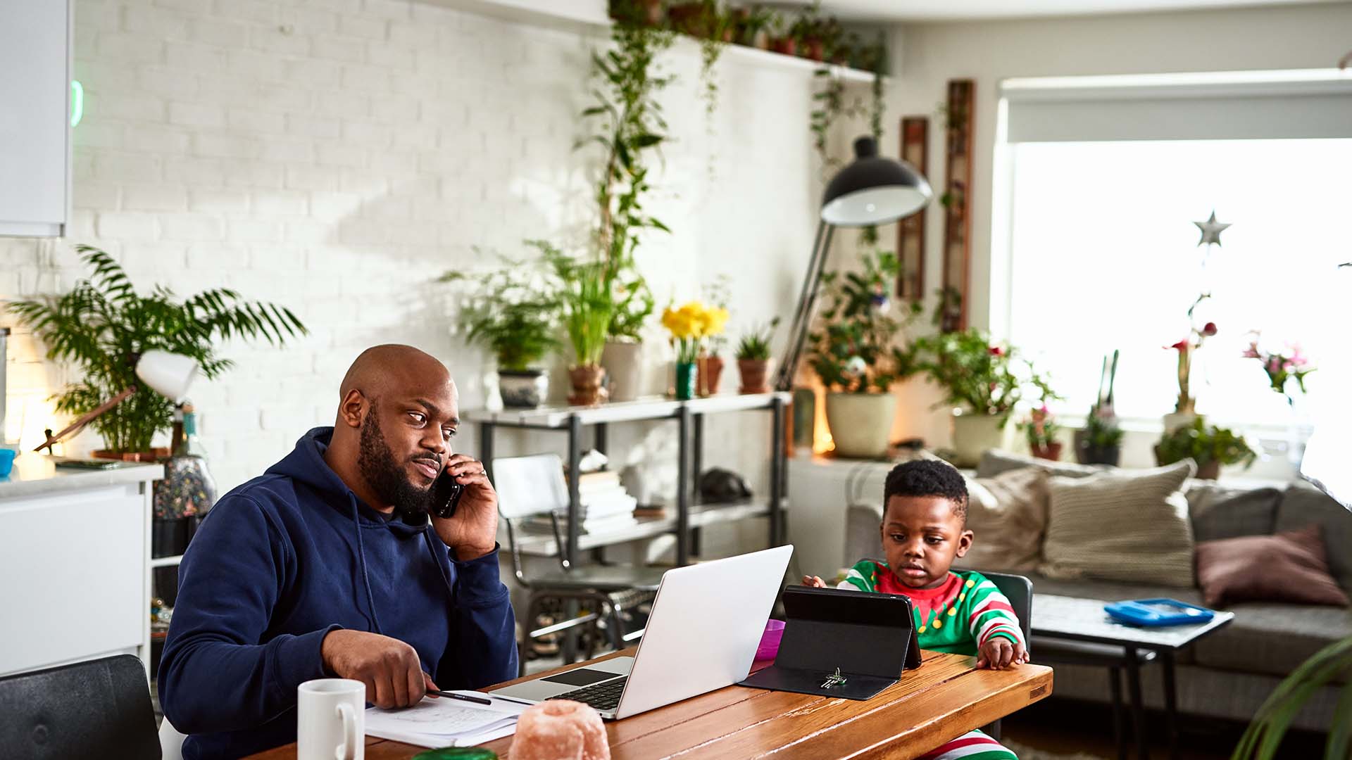 Mid adult man using phone at dining room table with laptop, boy sitting patiently with device, working from home, childcare issues, multi tasking.