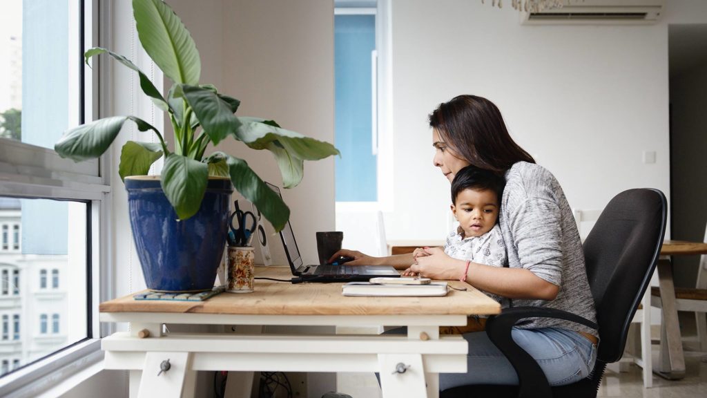 Woman holding baby looking at laptop screen on her desk.