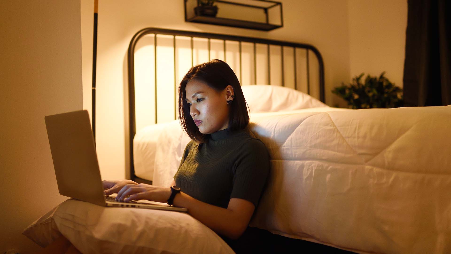 Woman sitting on floor leaning against bed on her laptop in low light