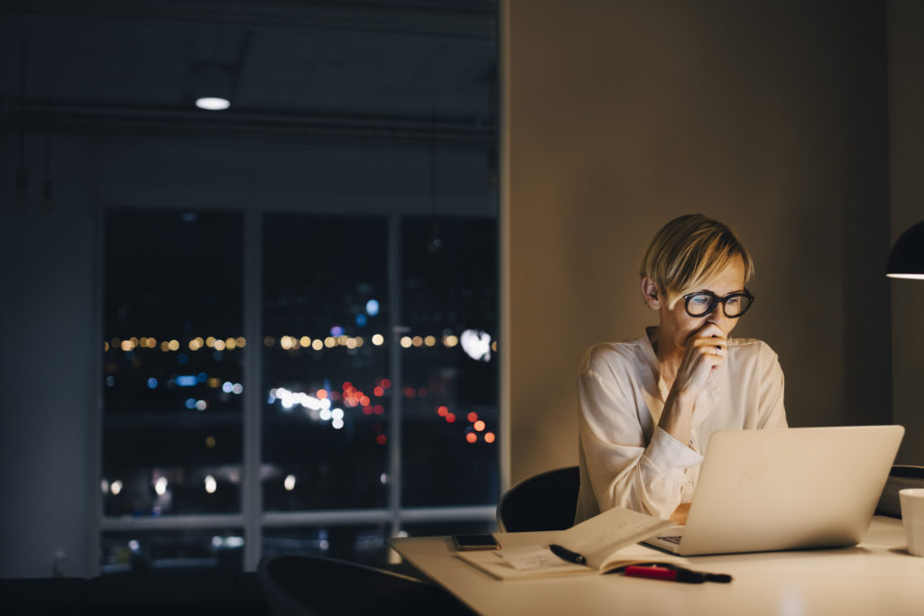 Woman sitting at desk staring at laptop screen with window of city in background