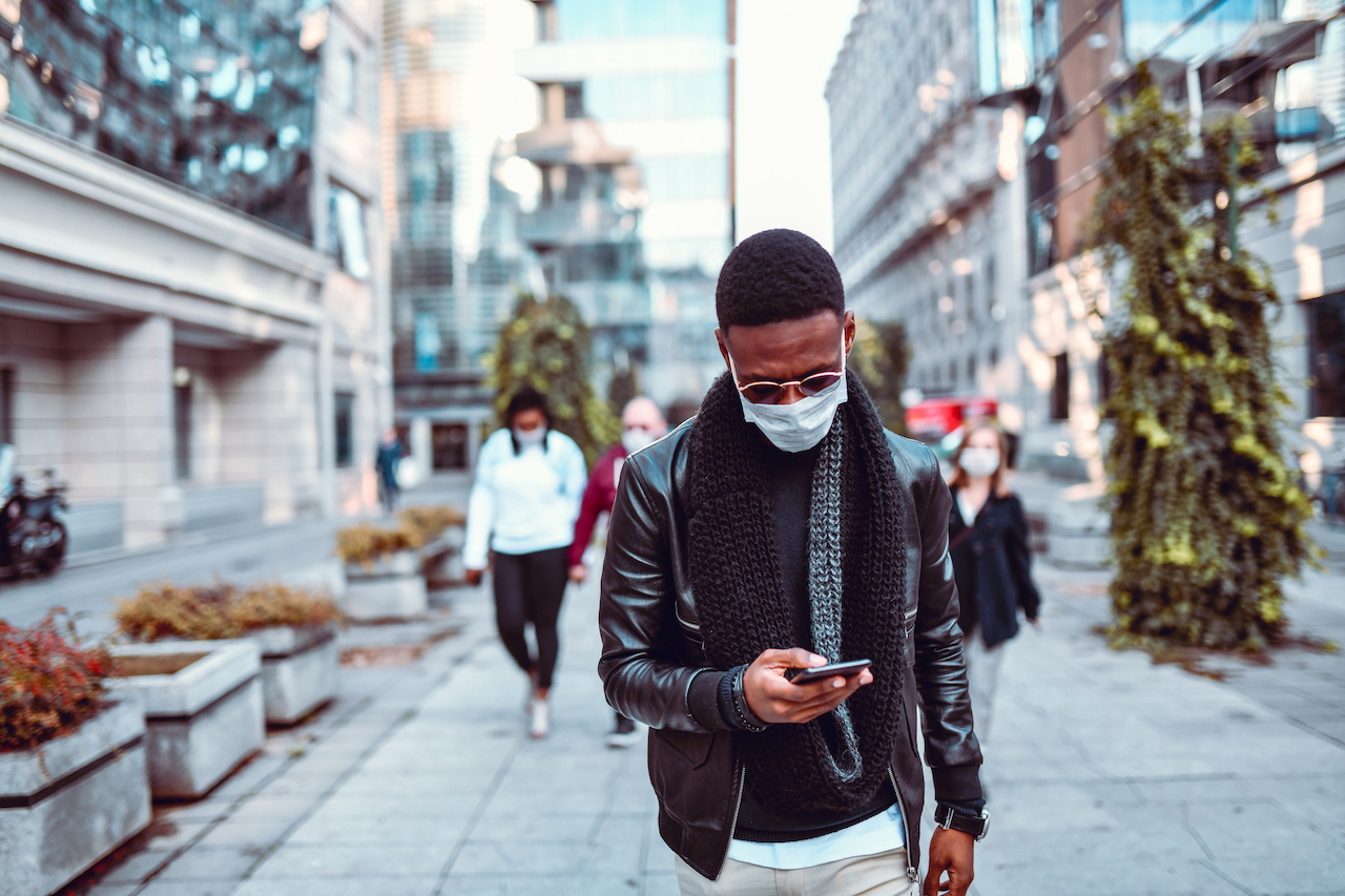 photo of a man walking through city street looking at his phone wearing a mask
