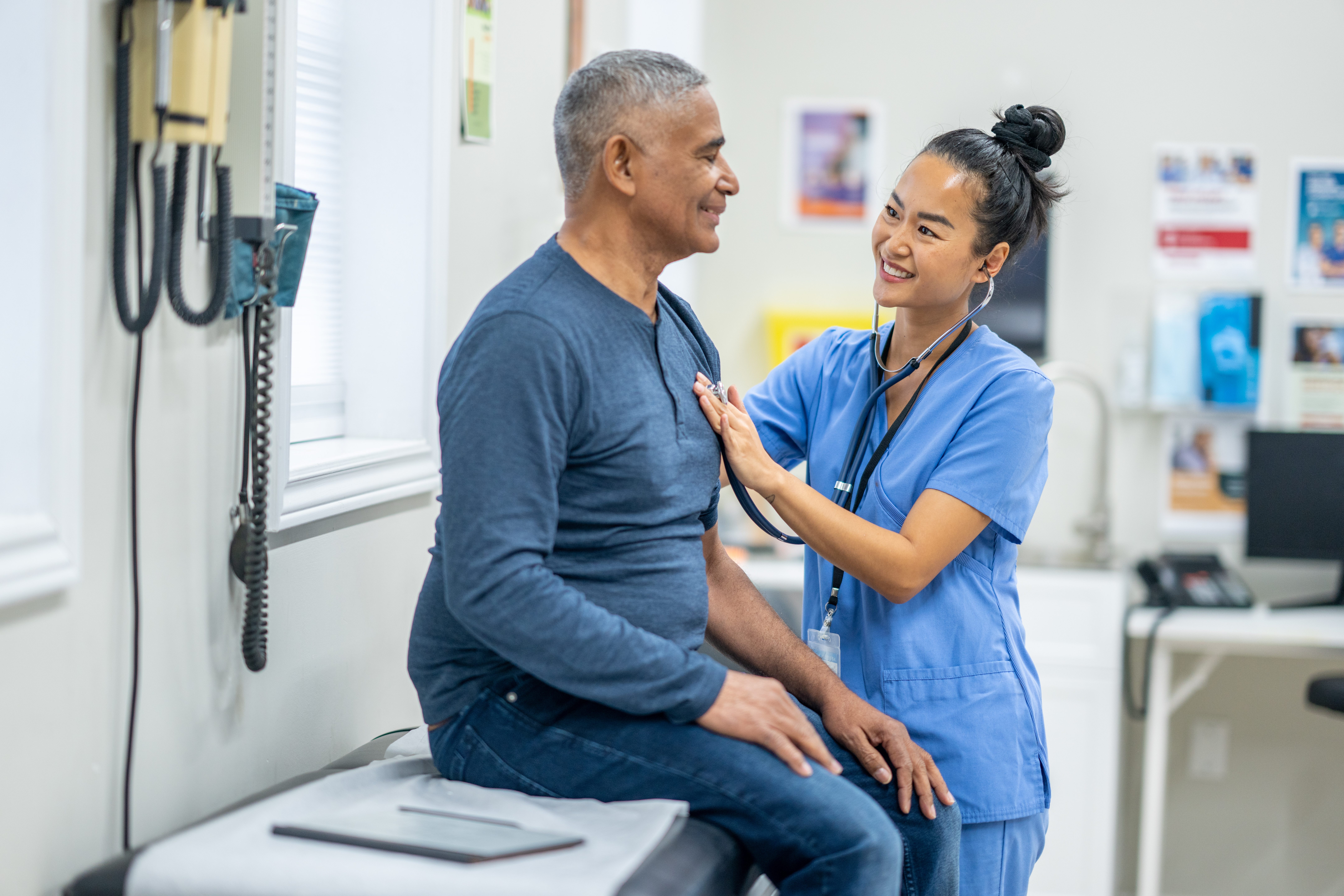 A senior gentleman of Hispanic decent, sits up on an exam table as his female nurse listens to his heart. The nurse is wearing blue scrubs and smiling up gently at the patient to put him at ease.